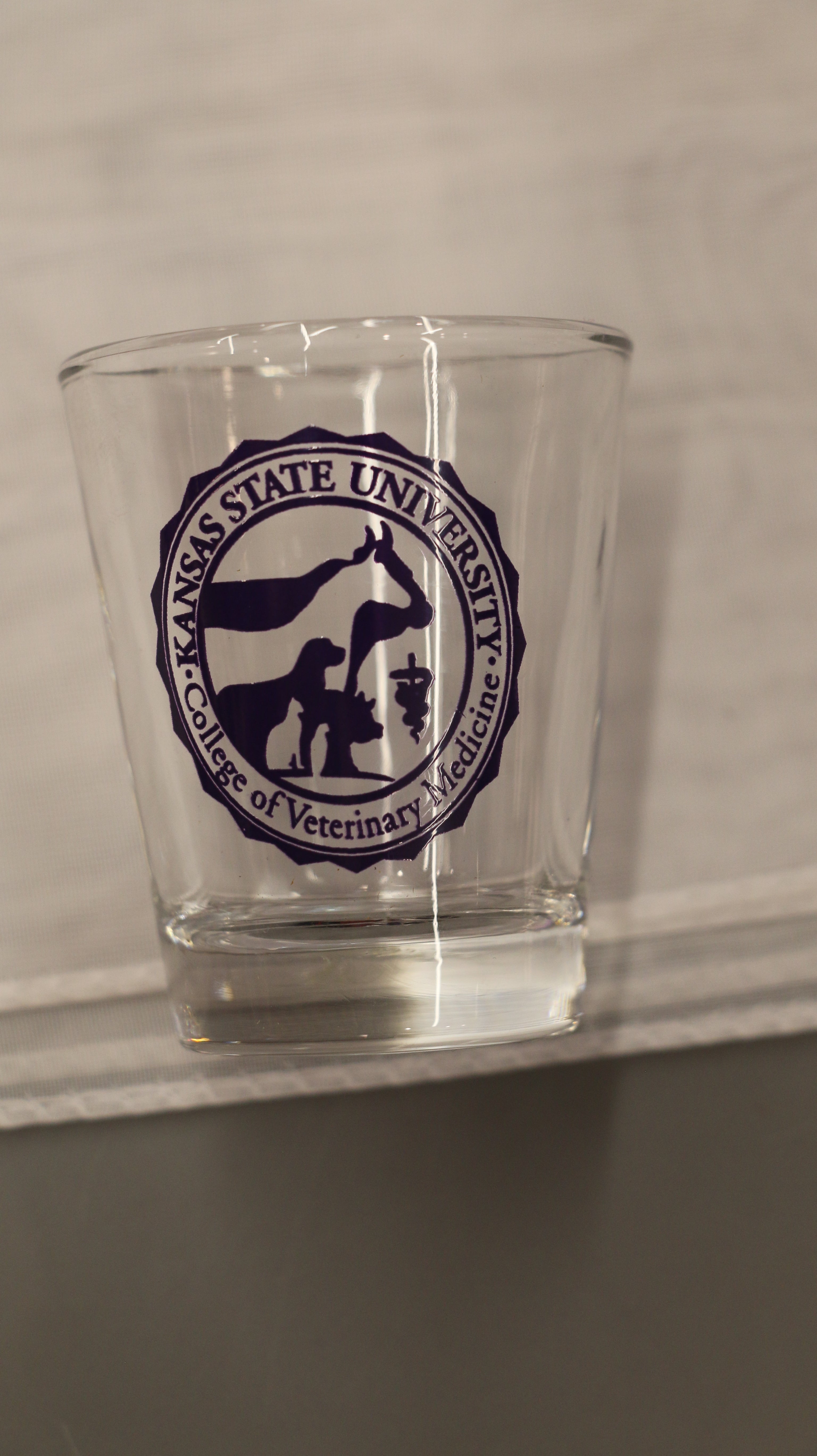 A clear 1.5oz shot glass with a purple custom CVM seal printed on it.