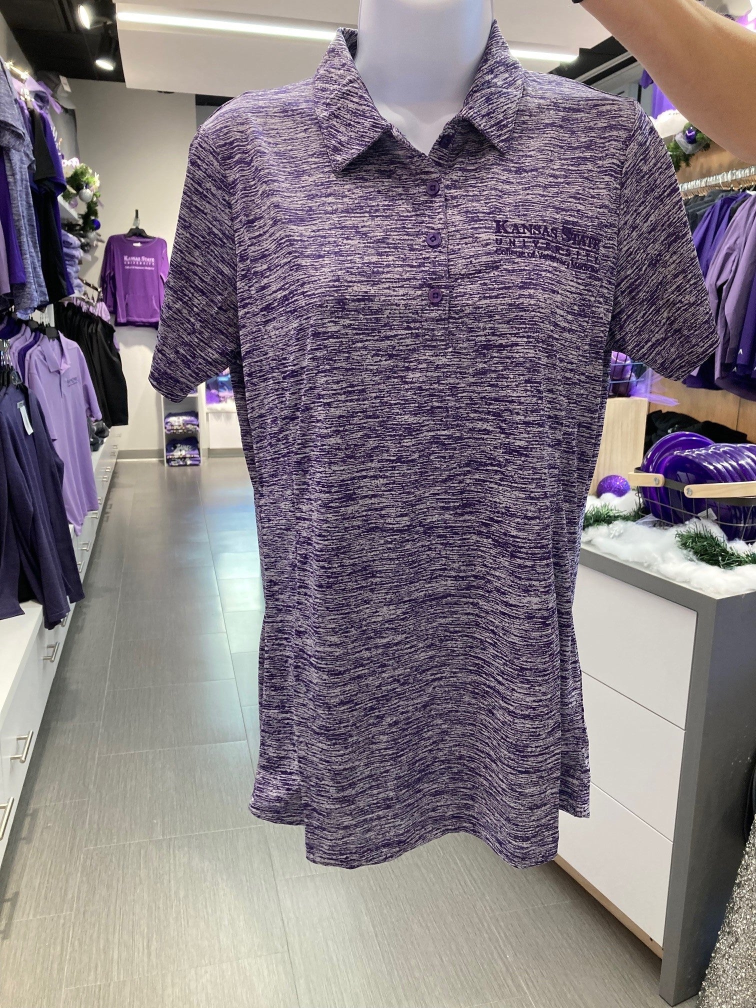 A purple and white dashed/striped patterned Sport-Tek ladies posicharge polo shirt displayed on a mannequin in a retail environment. There is a dark purple embroidered K-State CVM logo on the upper left chest area. 