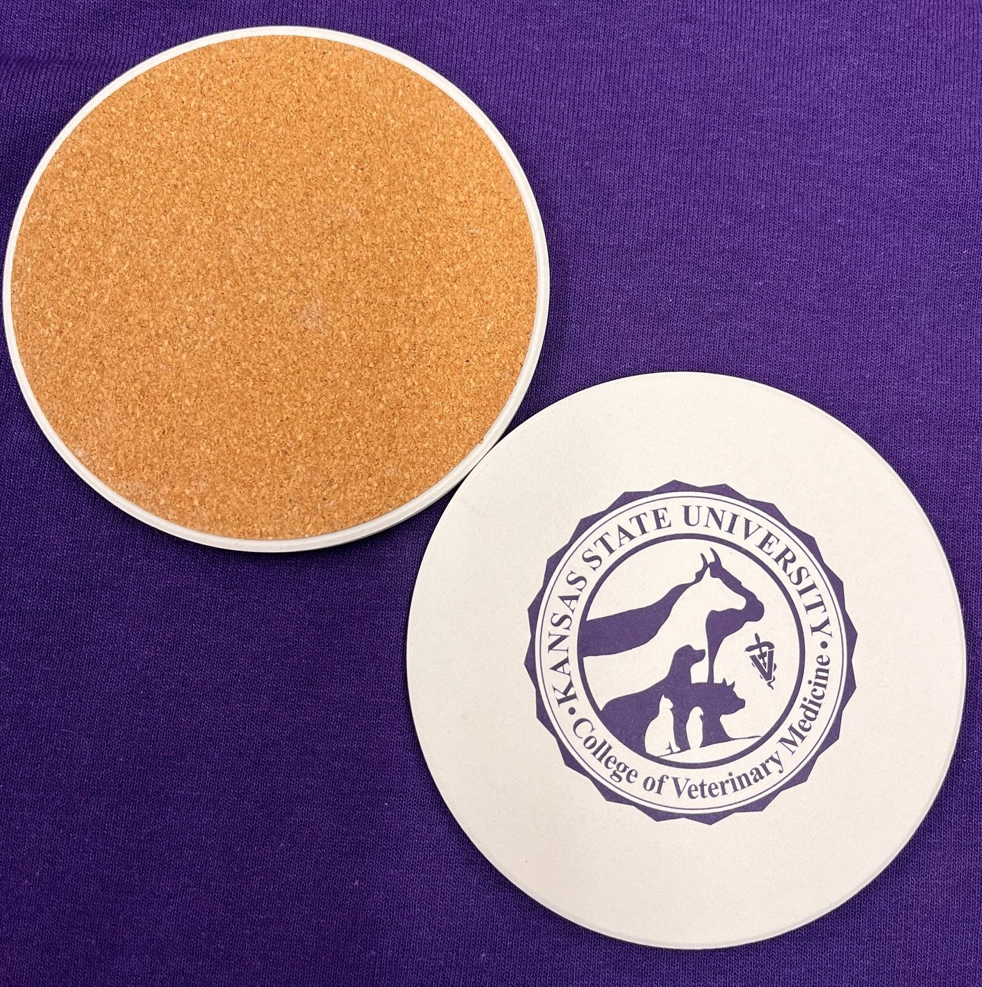 Round coaster with Kansas State University College of Veterinary Medicine seal in dark purple against a white background color. It also shows the cork textured underside of the coaster. 