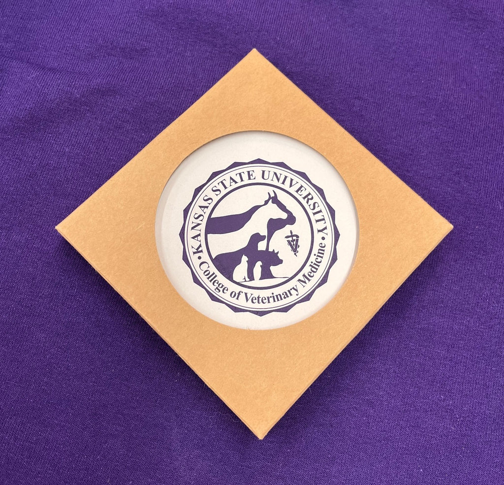 Round coaster with Kansas State University College of Veterinary Medicine seal in dark purple against a white background color and inside brown packaging. 