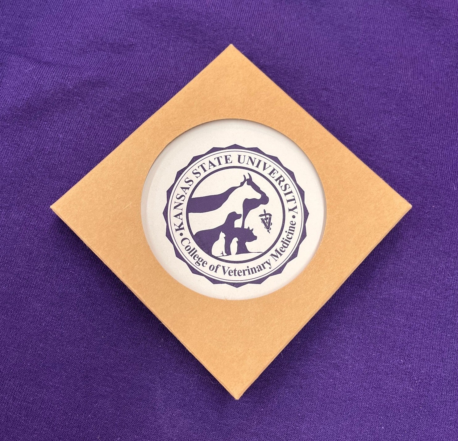 Round coaster with Kansas State University College of Veterinary Medicine seal in dark purple against a white background color and inside brown packaging. 