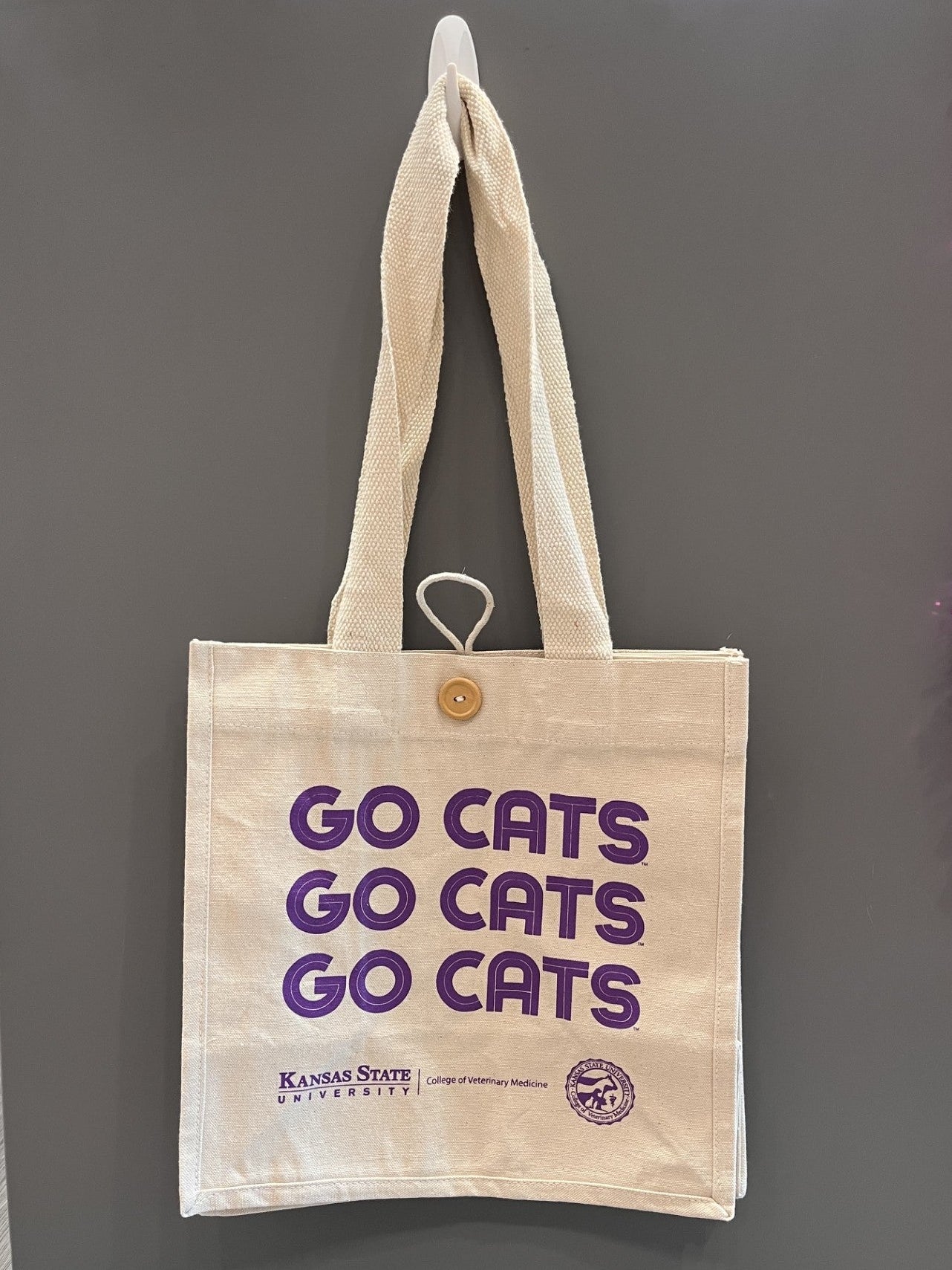 A cream canvas tote bag with purple 'GO CATS' text and a small Kansas State University logo, hanging on a gray display stand.