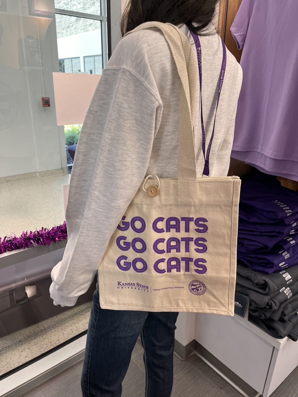A person wearing a light gray sweatshirt and dark jeans carries a beige canvas tote bag with purple 'GO CATS' text and Kansas State University branding in a retail store setting.