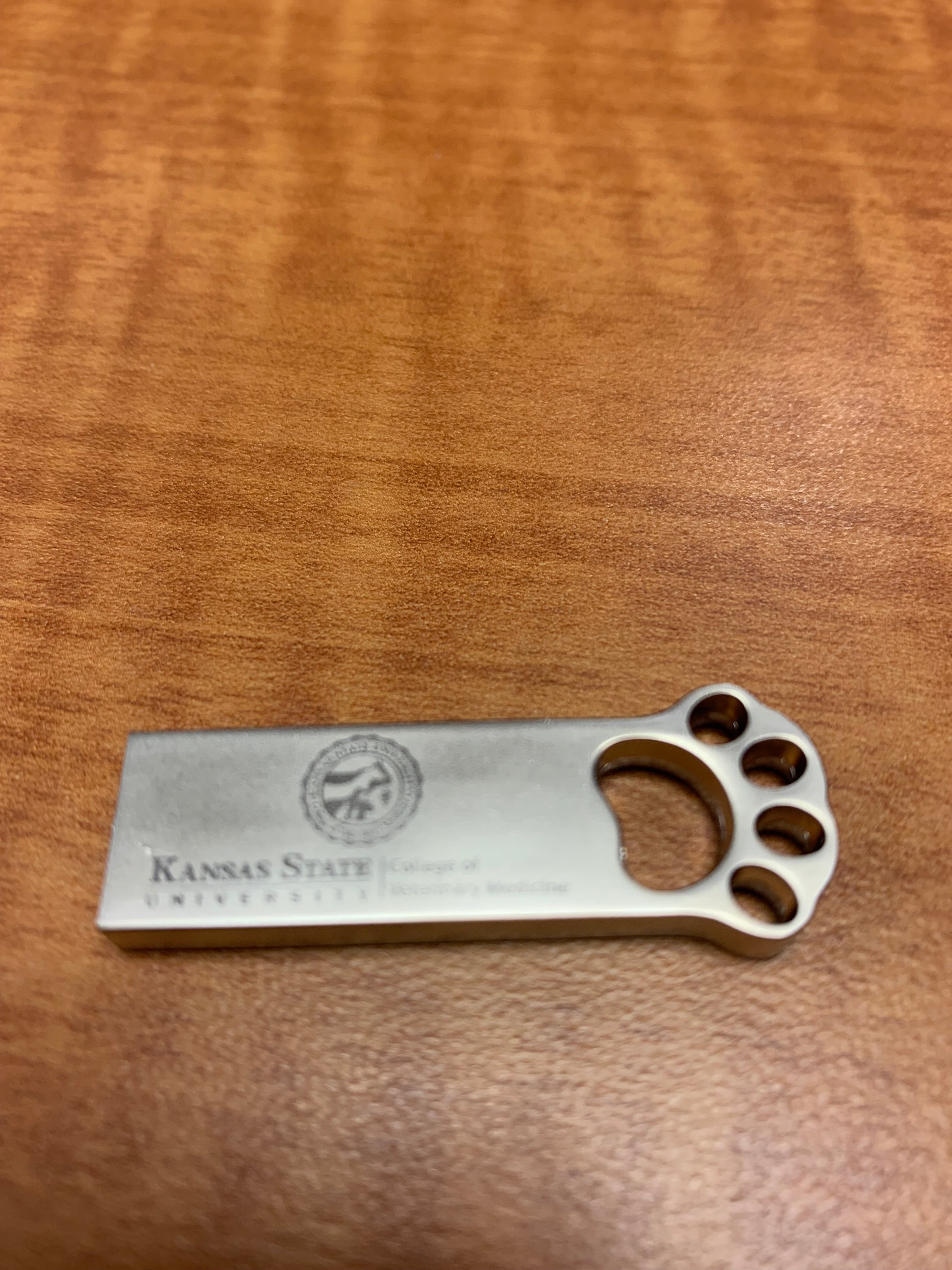 A silver USB flash drive with a pawprint design and K-State CVM logo engraved on it.