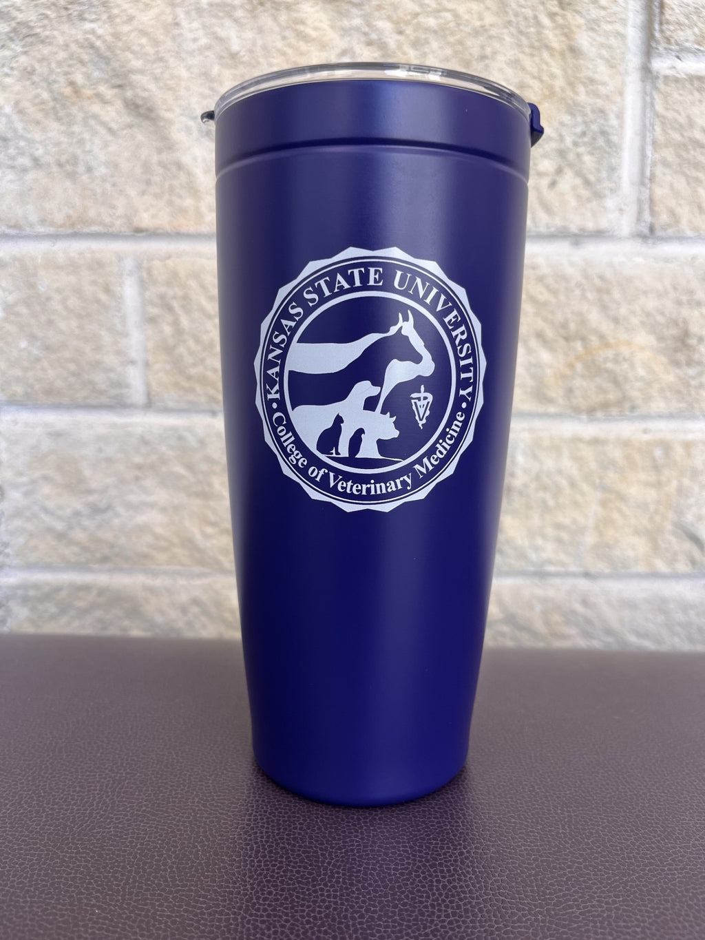 A purple insulated travel tumbler with a clear lid, featuring a white printed college of veterinary medicine seal, set against a light stone wall background