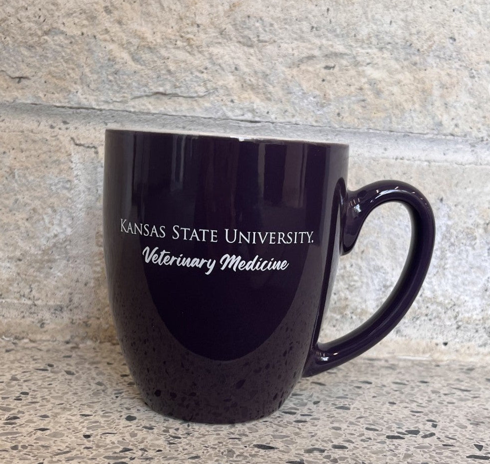A dark purple ceramic coffee mug with 'KANSAS STATE UNIVERSITY Veterinary Medicine' printed in white, sitting on a speckled stone countertop against a stone wall.