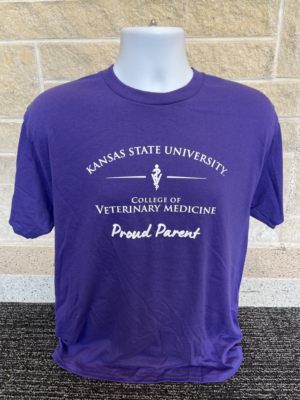 A purple short-sleeve t-shirt displayed on a white mannequin torso against a light tan brick wall and dark textured carpet. The shirt features a white printed graphic that reads 'KANSAS STATE UNIVERSITY.