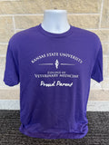 A purple short-sleeve t-shirt displayed on a white mannequin torso against a light tan brick wall and dark textured carpet. The shirt features a white printed graphic that reads 'KANSAS STATE UNIVERSITY.