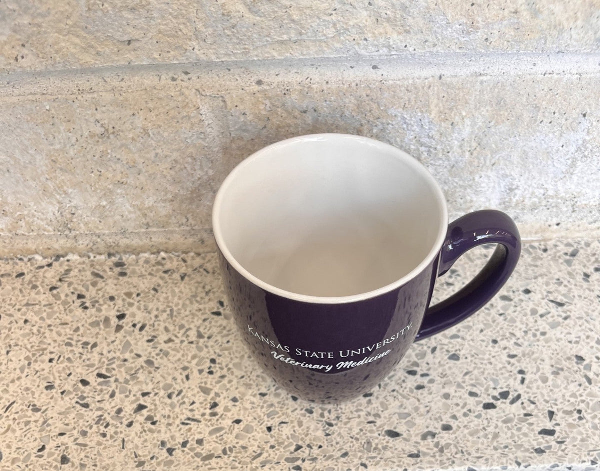 A dark purple ceramic coffee mug with a white interior and handle, featuring the text 'KANSAS STATE UNIVERSITY' and 'Veterinary Medicine' printed on the front. The mug is placed on a speckled stone countertop with a dark textured rug below.