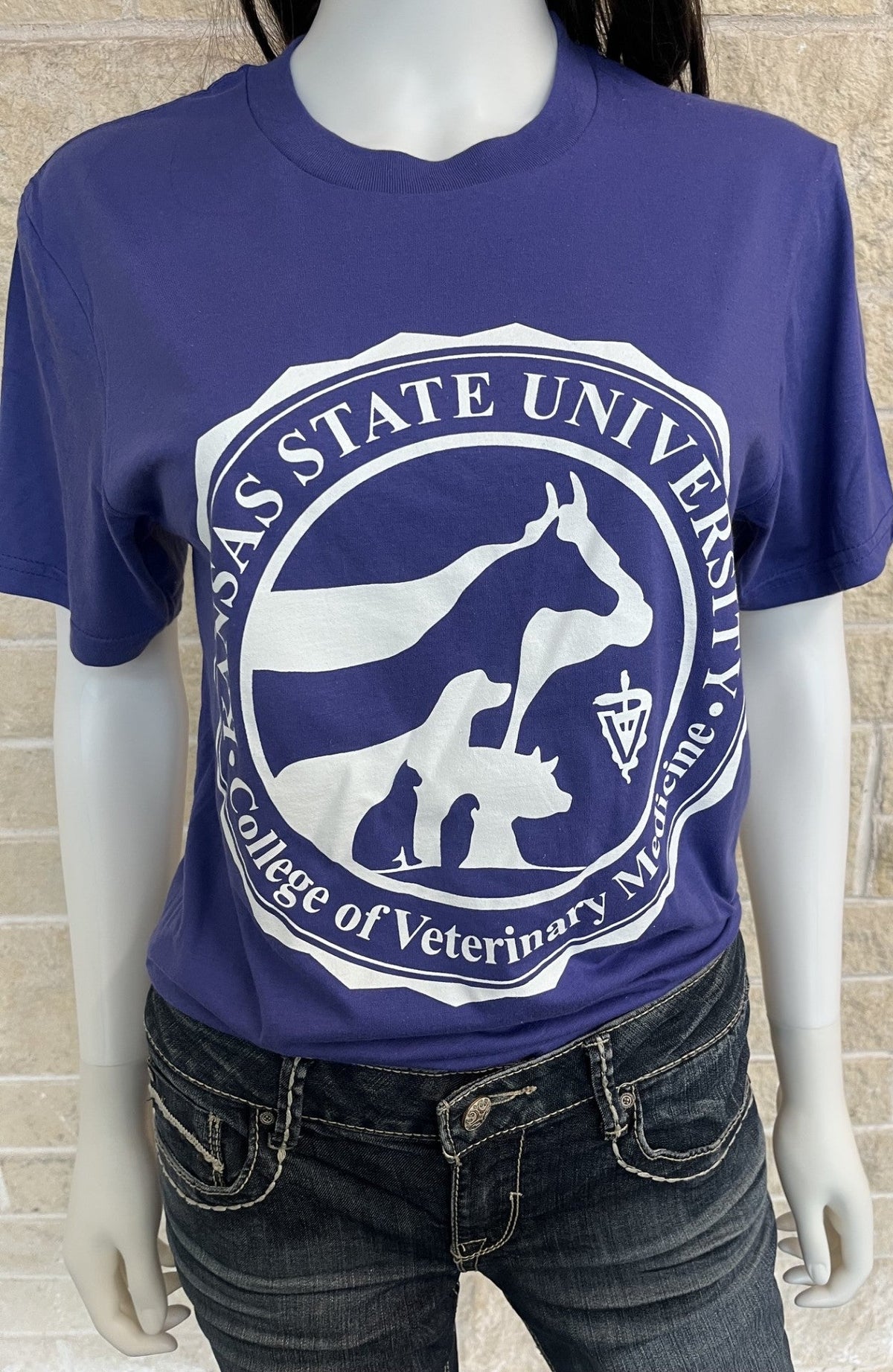 A purple short-sleeve t-shirt with a large white Kansas State University College of Veterinary Medicine seal printed on the front, displayed on a mannequin wearing dark wash jeans against a light tan brick wall.