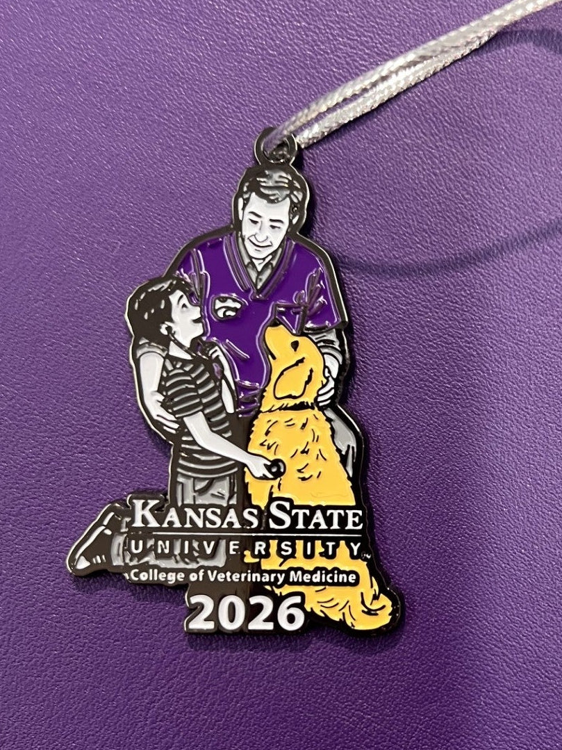 A close-up of a Christmas Ornament with  a silver cord. A little boy with a Veterinarian and a yellow lab with Kansas State University College of Veterinary Medicine logo with the year 2026 in white. 