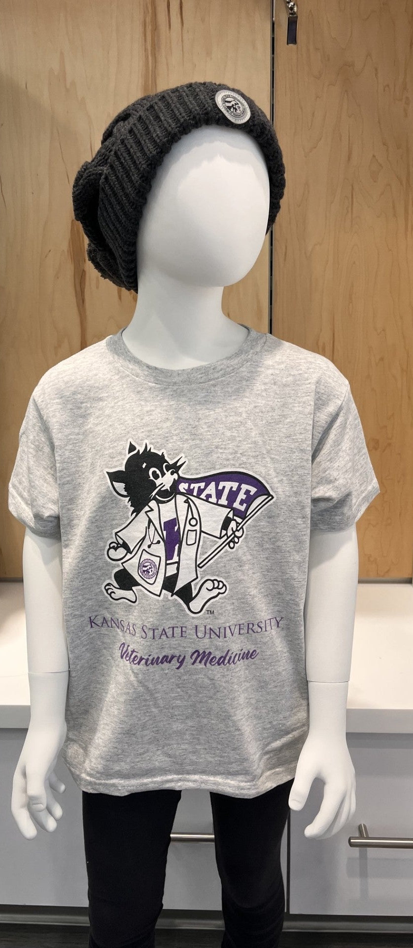 A mannequin wearing a light gray heather graphic T-shirt with Lab Coat Willie and 'Kansas State University Veterinary Medicine' print, paired with a black knit beanie and black pants, displayed in a retail store setting.