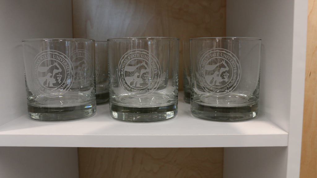 Multiple clear old fashion glasses with the CVM seal logos in the center on a shelf. 