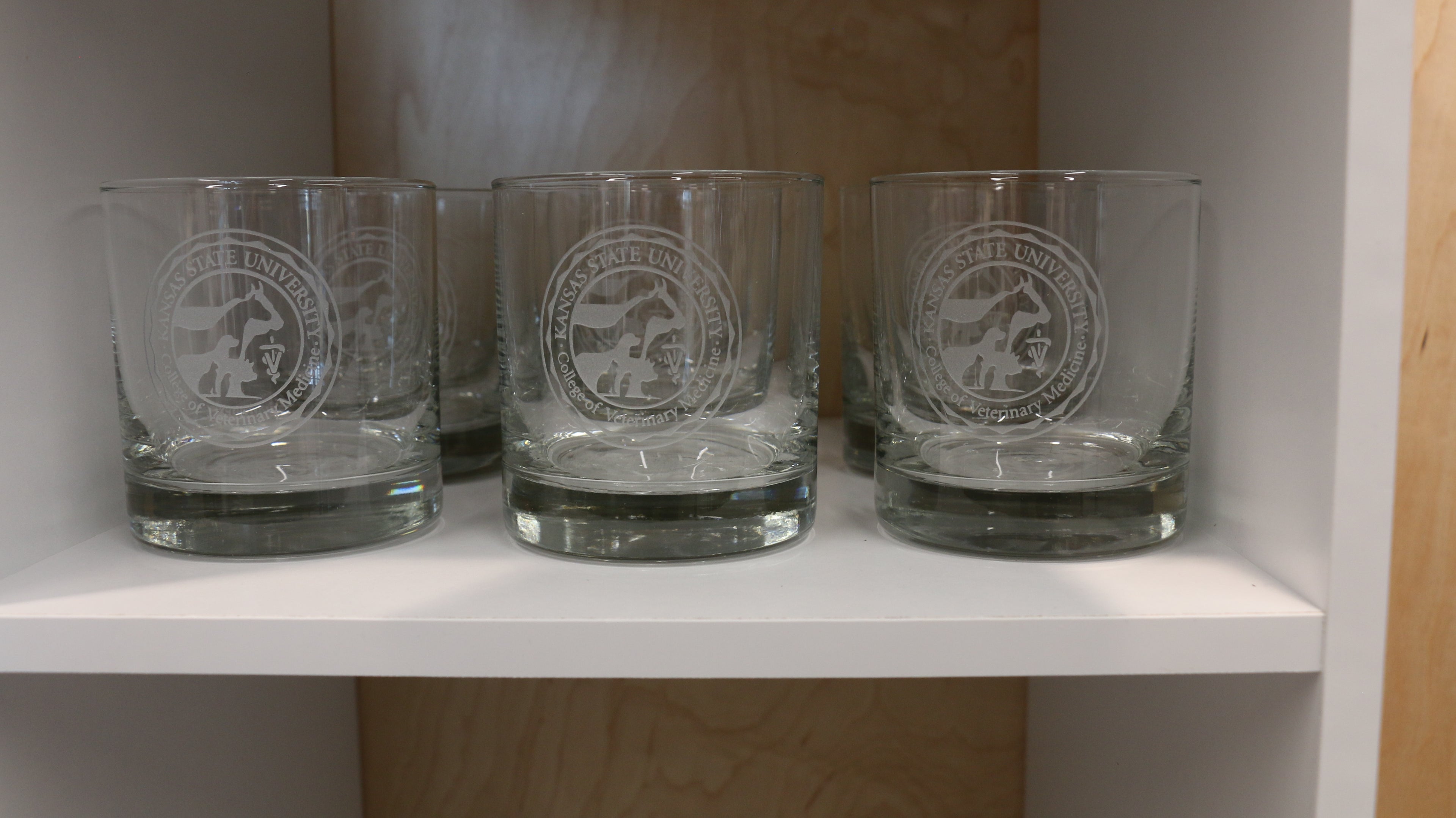 Multiple clear old fashion glasses with the CVM seal logos in the center on a shelf. 