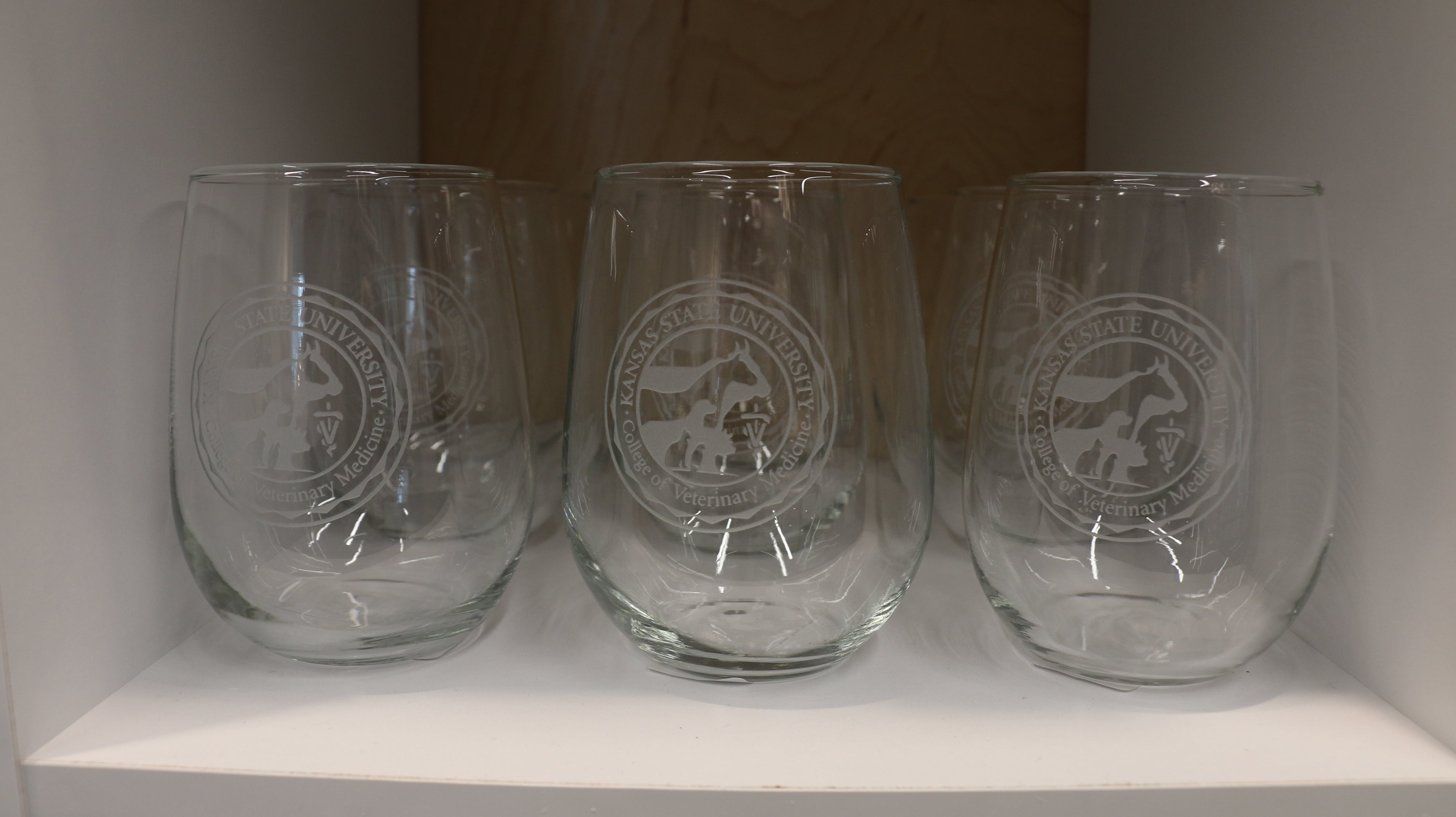 Multiple clear stemless wine glasses with etched CVM seals in the center on a shelf. 