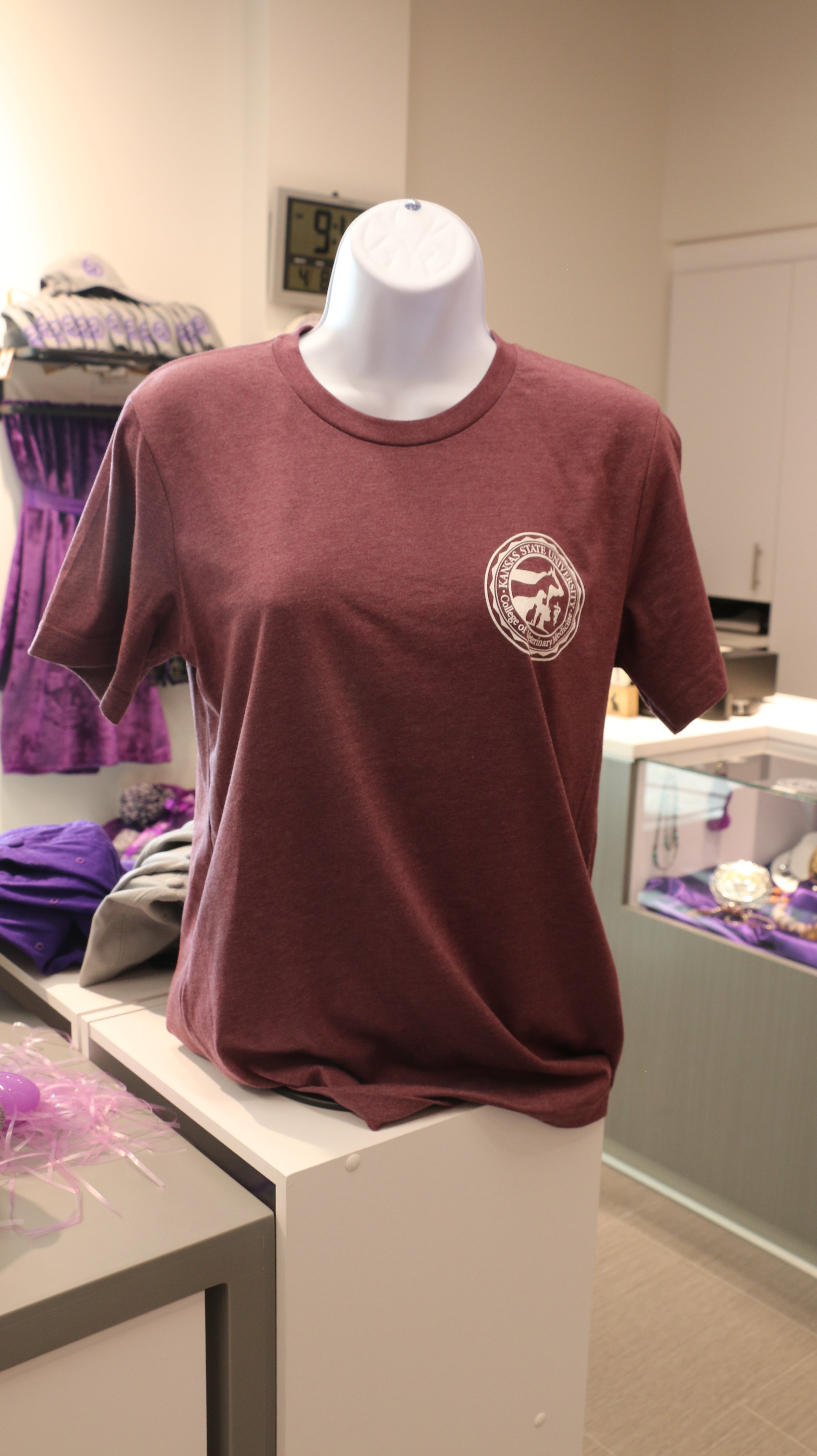 A maroon Bella Canvas V-neck t-shirt with a white CVM seal on the upper left chest, displayed on a mannequin in a retail setting.