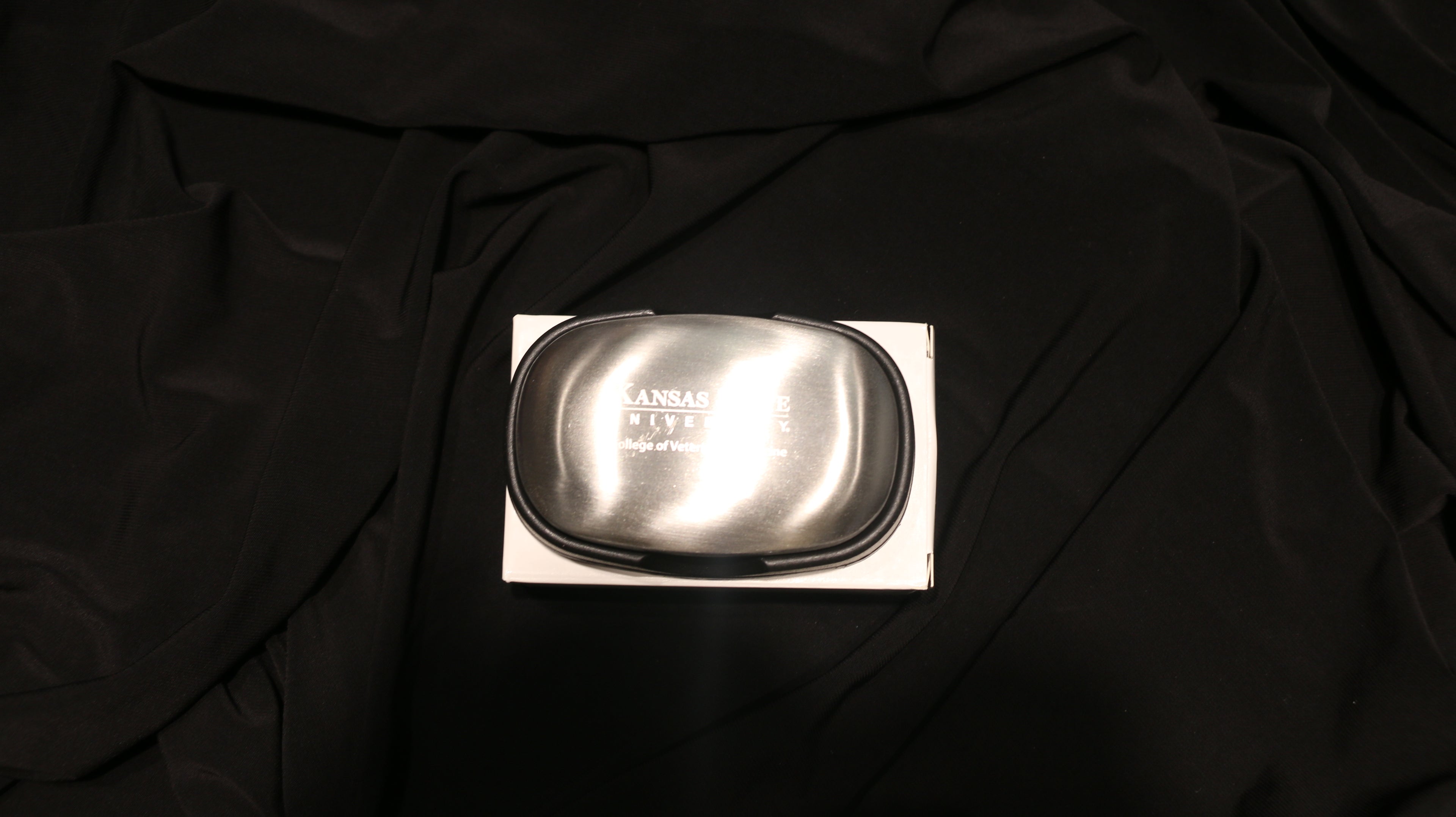 A silver steel soap bar on a black background, packaged in a cardboard box with the CVM logo engraved in the center. 