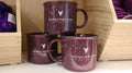 A set of three purple ceramic campfire mugs with white speckled patterns and the Veterinary Health Center logo printed on them, displayed on a shelf.