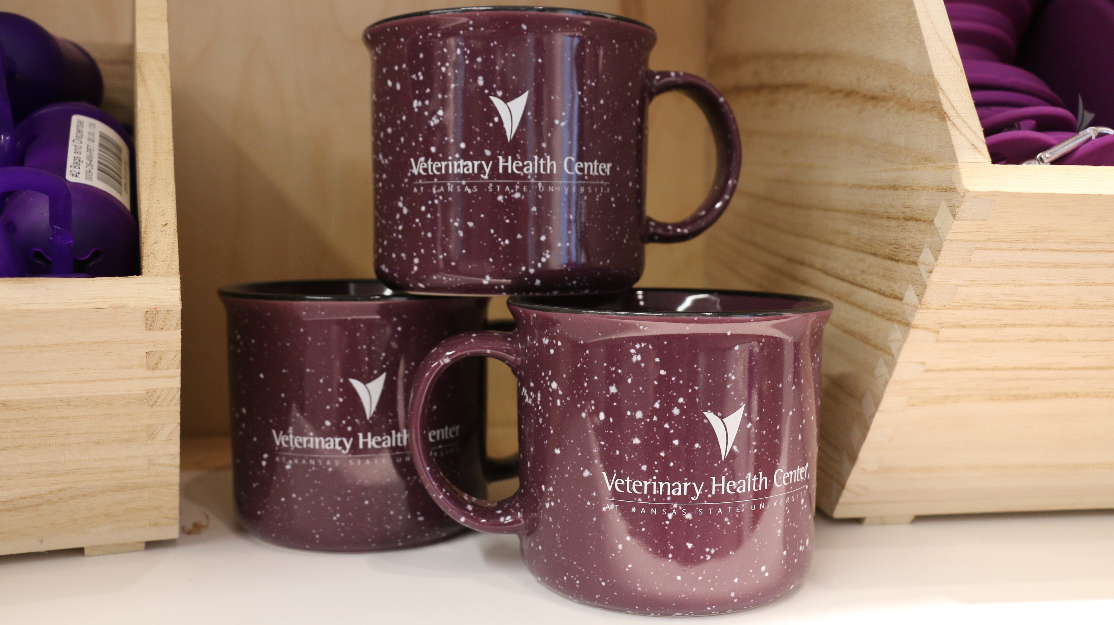 A set of three purple ceramic campfire mugs with white speckled patterns and the Veterinary Health Center logo printed on them, displayed on a shelf.
