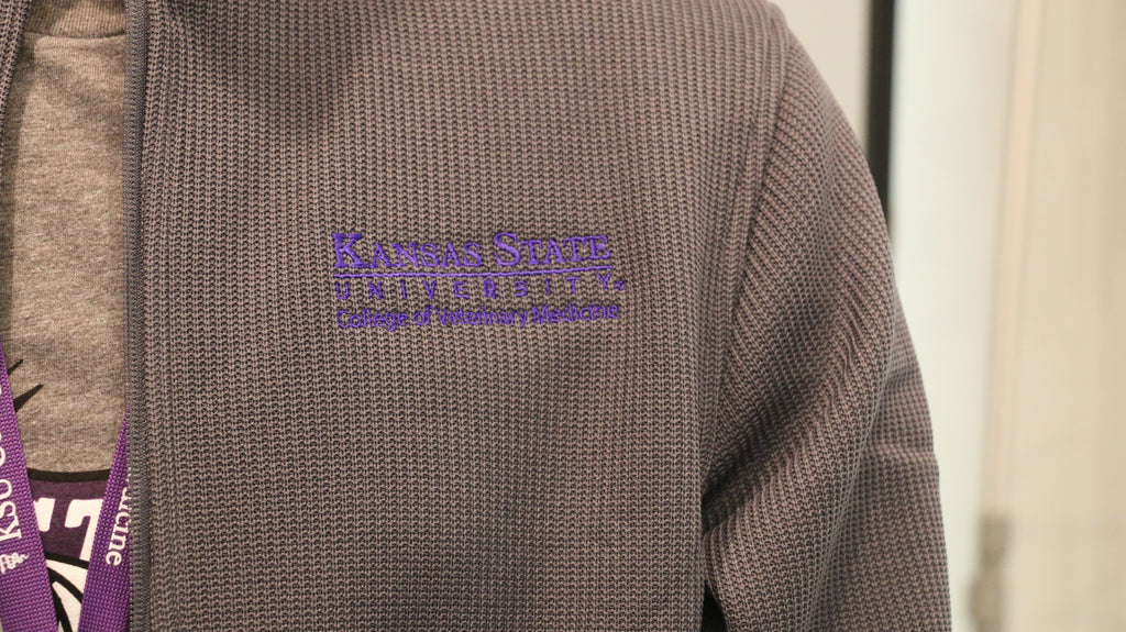 A close-up view of the purple embroidered Kansas State University College of Veterinary Medicine logo on the upper left chest area. 