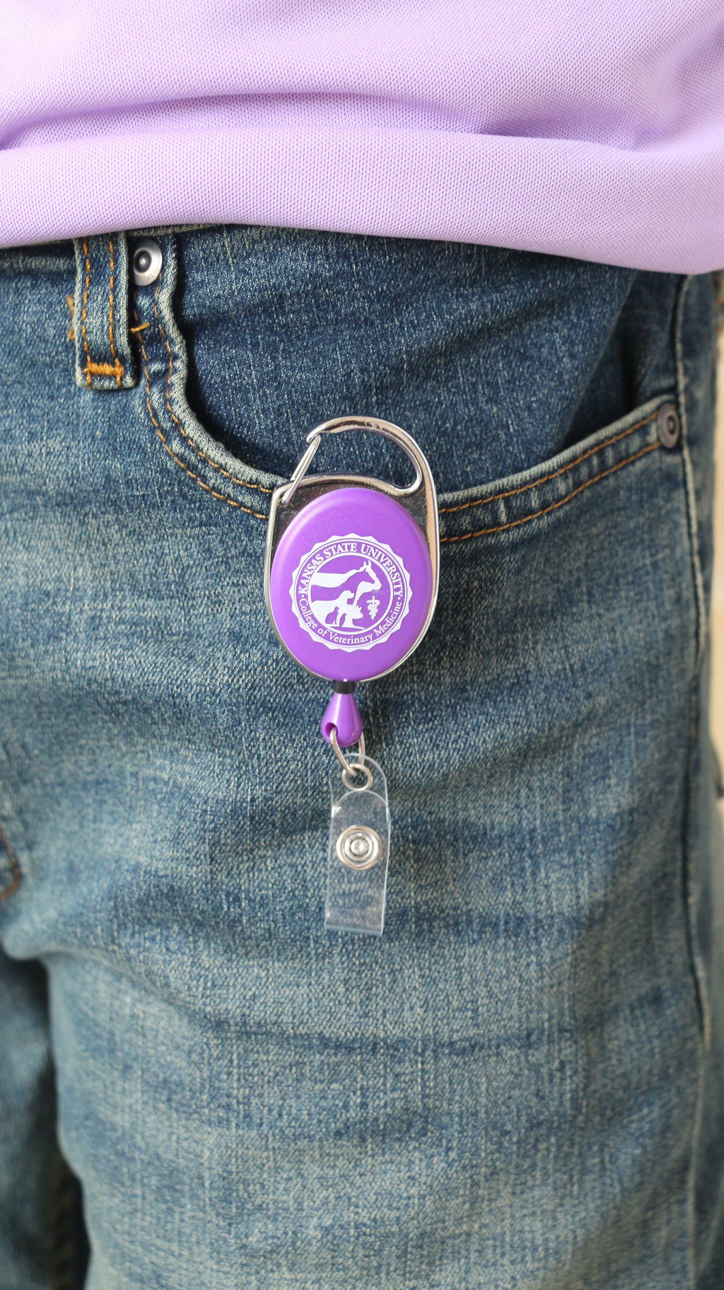 A clip-on retractable badge holder in purple and chrome color attached to a pair of jeans with the CVM seal in the center.
