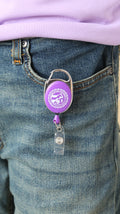 A clip-on retractable badge holder in purple and chrome color attached to a pair of jeans with the CVM seal in the center.
