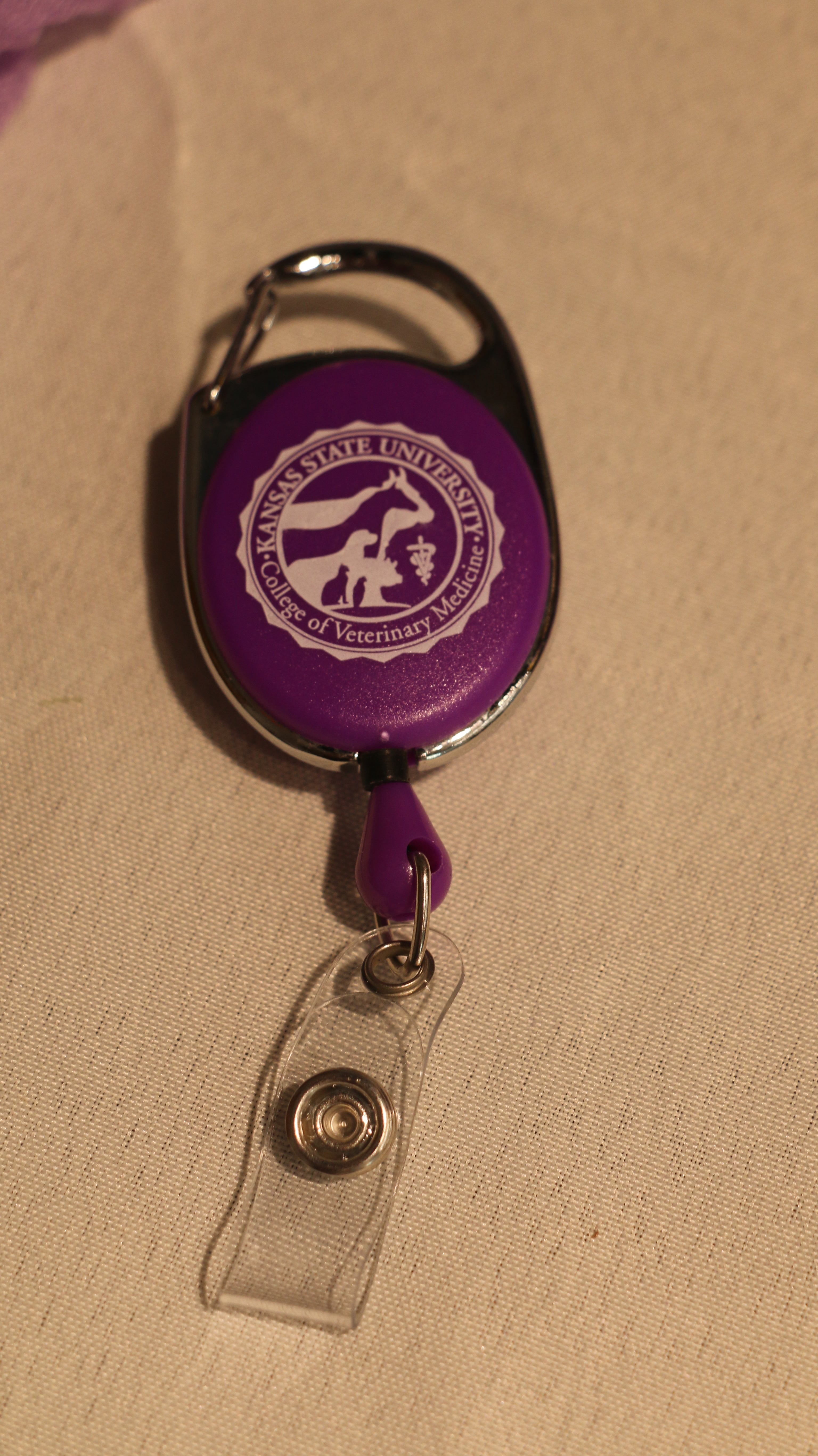 A clip-on retractable badge holder in purple and chrome color attached to a pair of jeans with the CVM seal in the center.