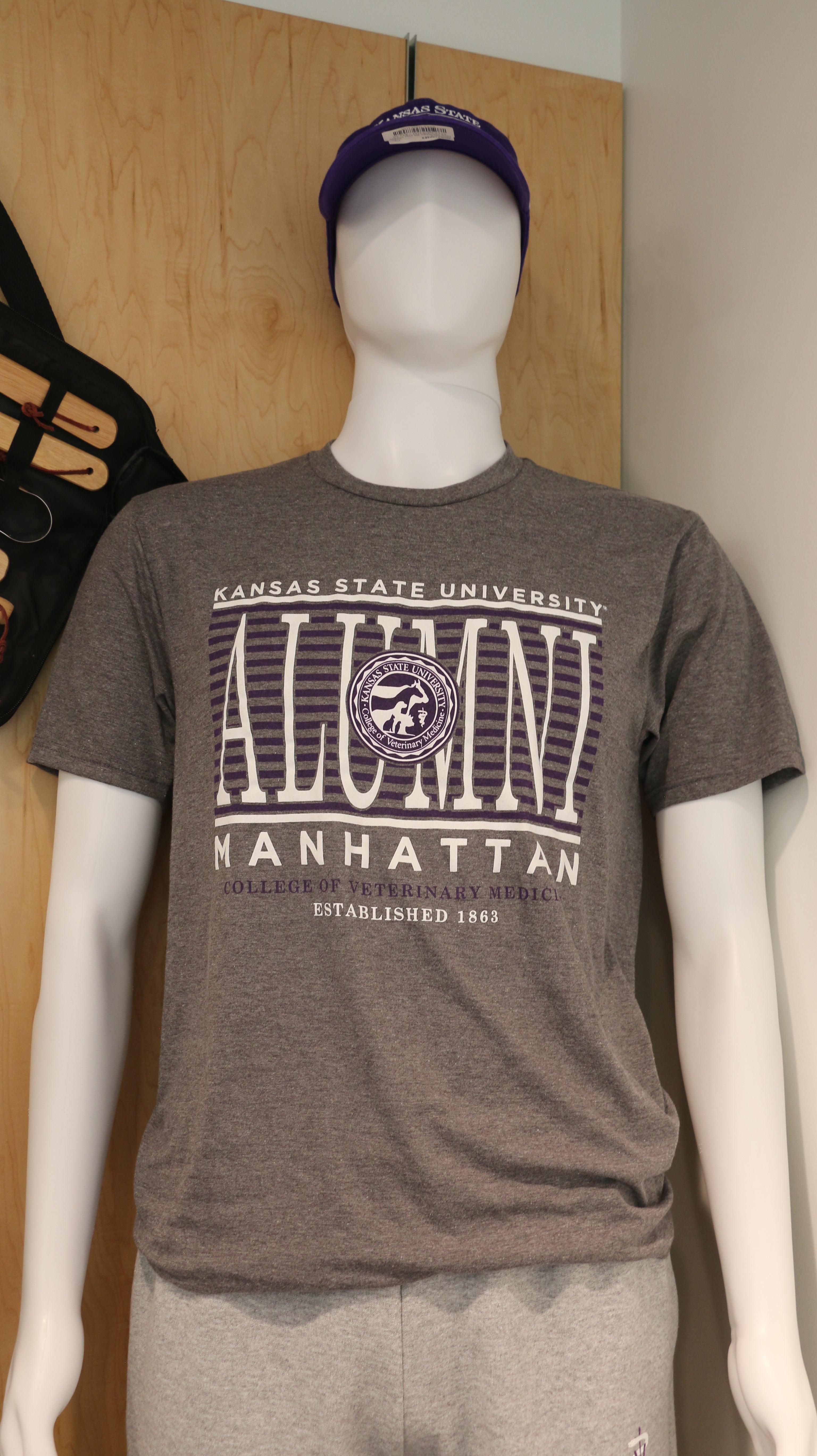 A mannequin wearing a grey District Alumni T-Shirt with text 'Alumni Manhattan' on the front with a purple and white CVM seal. 