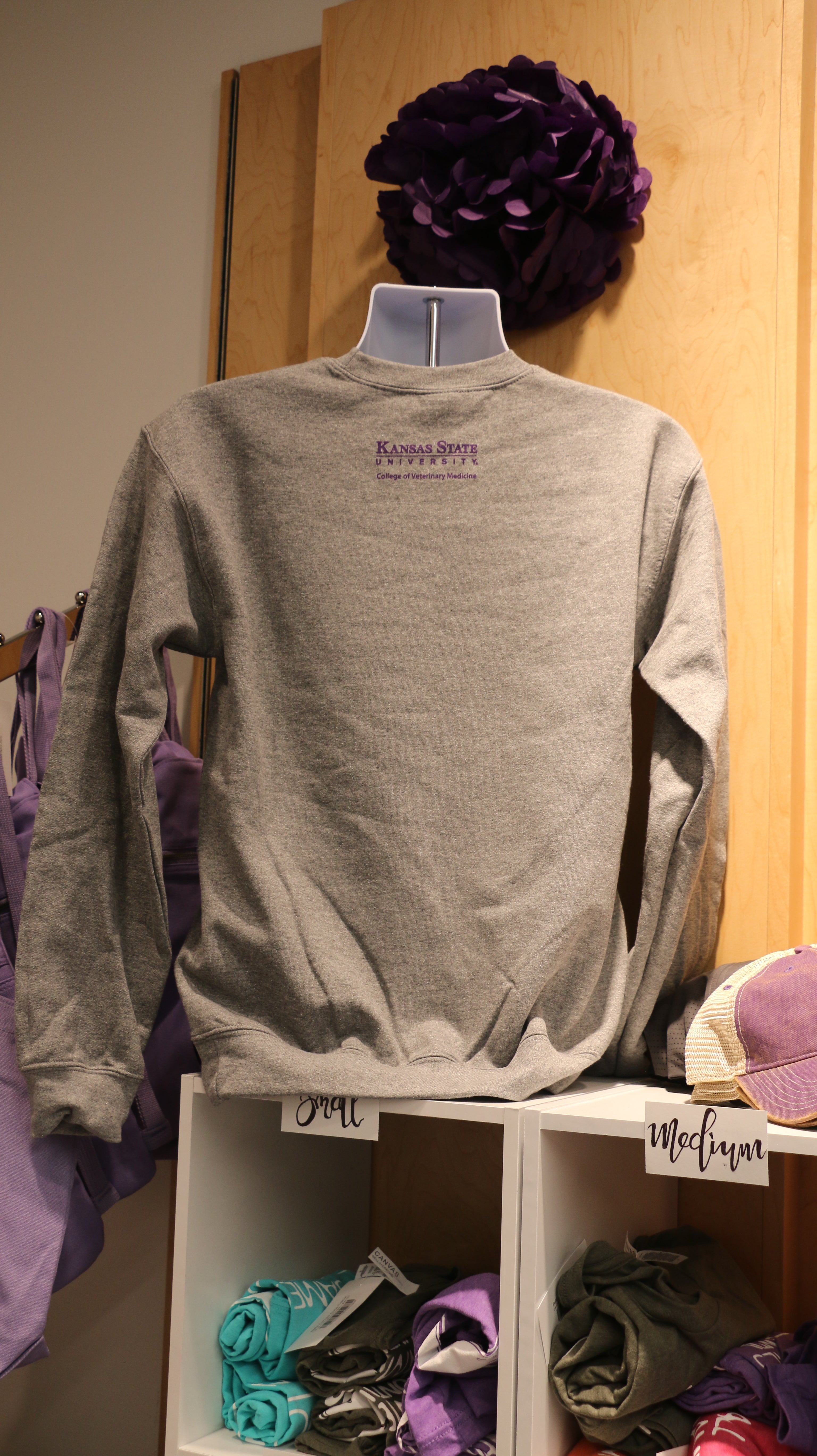 A rear view of the grey crewneck sweatshirt with a purple CVM logo on the upper middle back area. 
