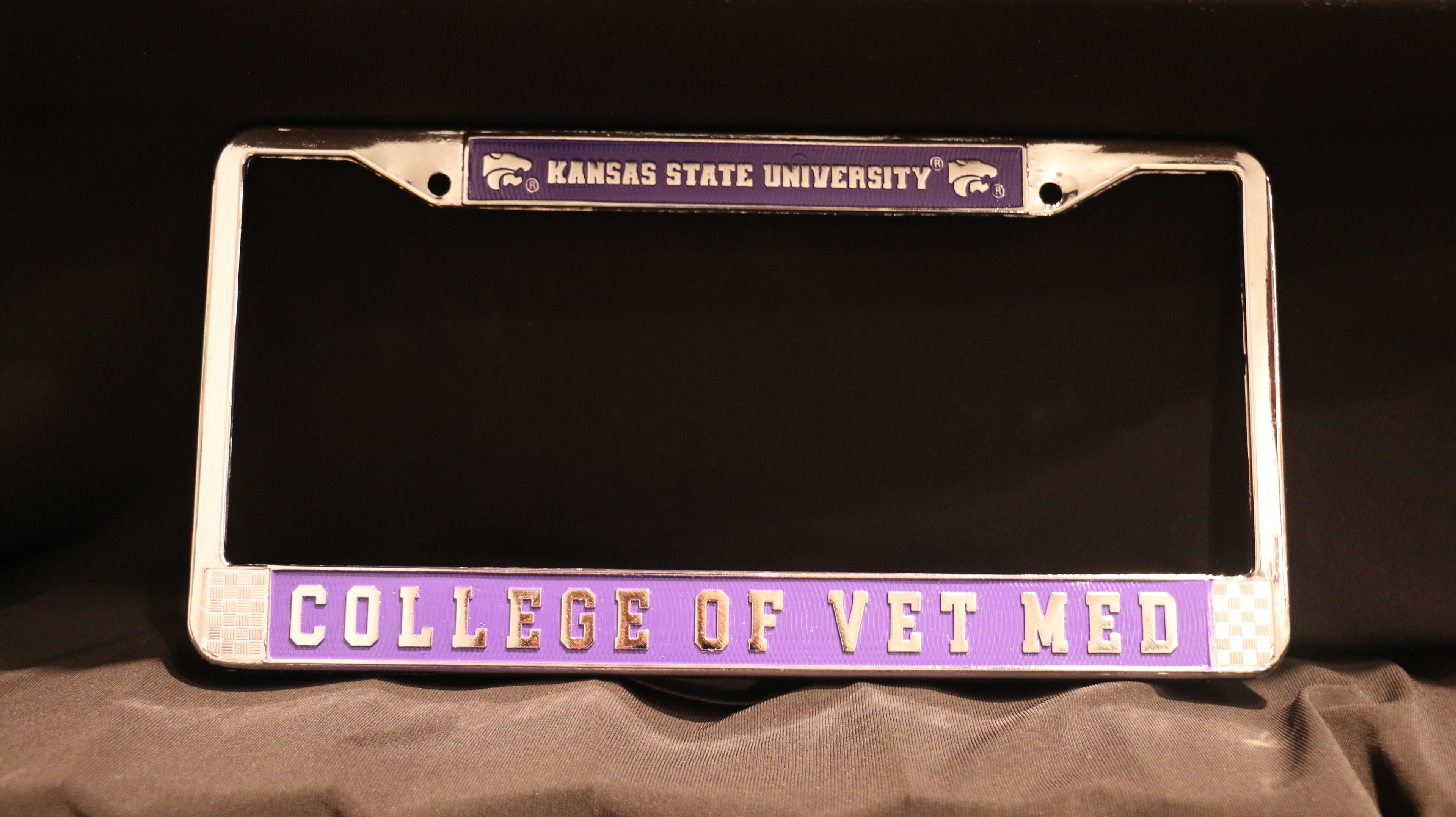 A metal license plate holder with 'Kansas State University' and 'College of Vet Med' embossed on it, in silver and purple color scheme.