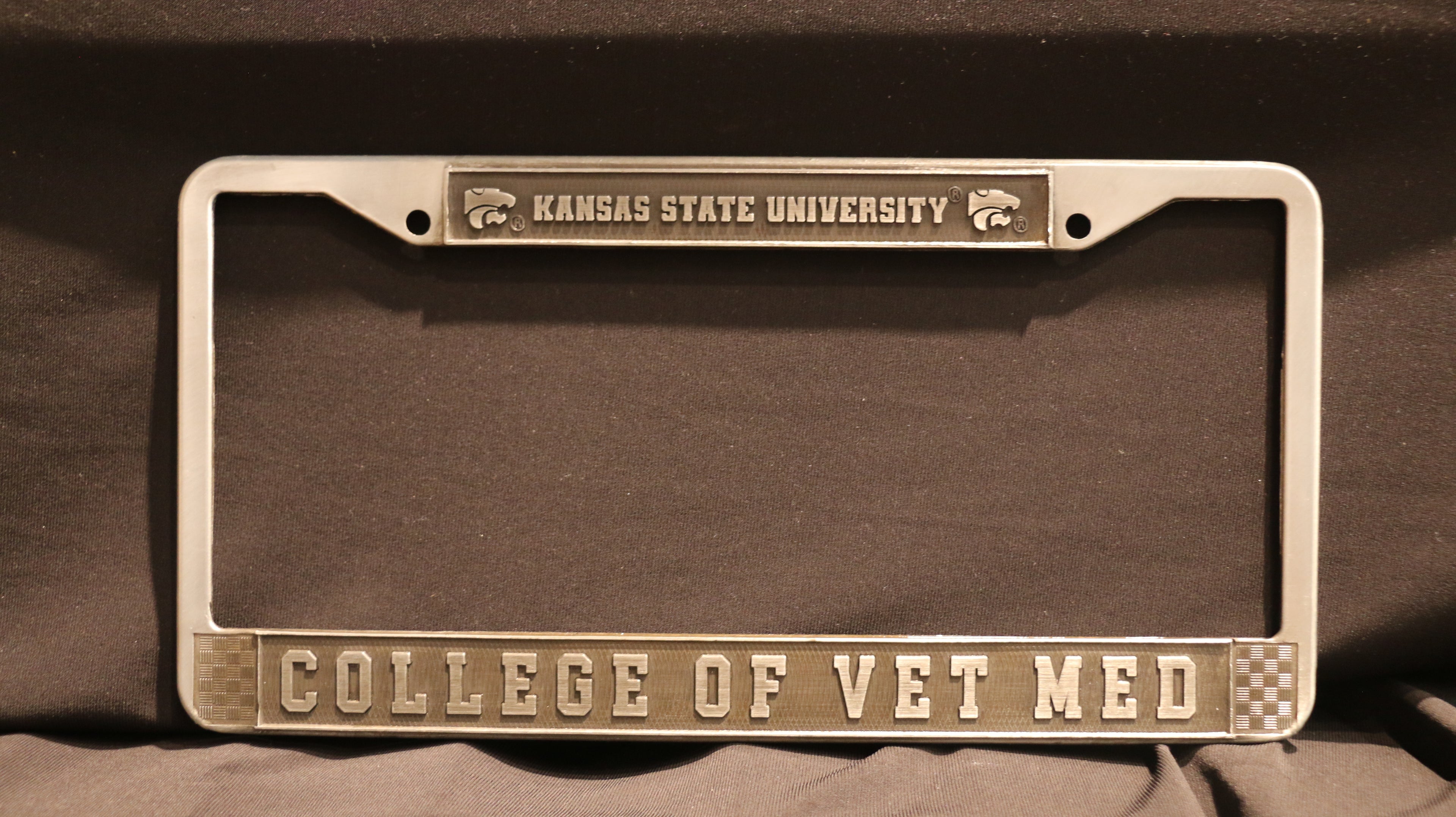 A metal license plate holder with 'Kansas State University' and 'College of Vet Med' embossed on it, in silver and grey color scheme.