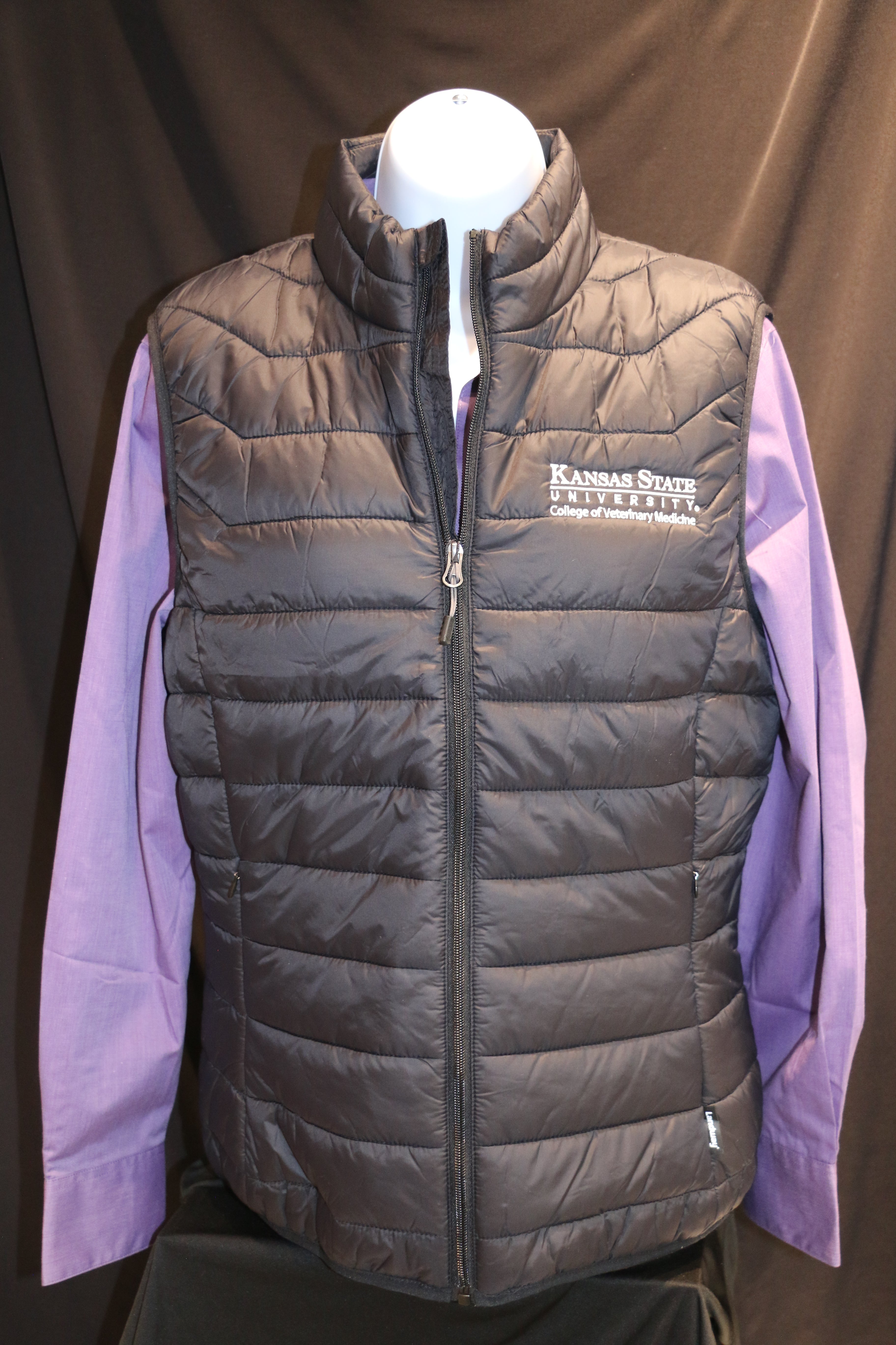 A black quilted puffer vest with zippered pockets, white K-State logo, and a stand-up collar on a mannequin.
