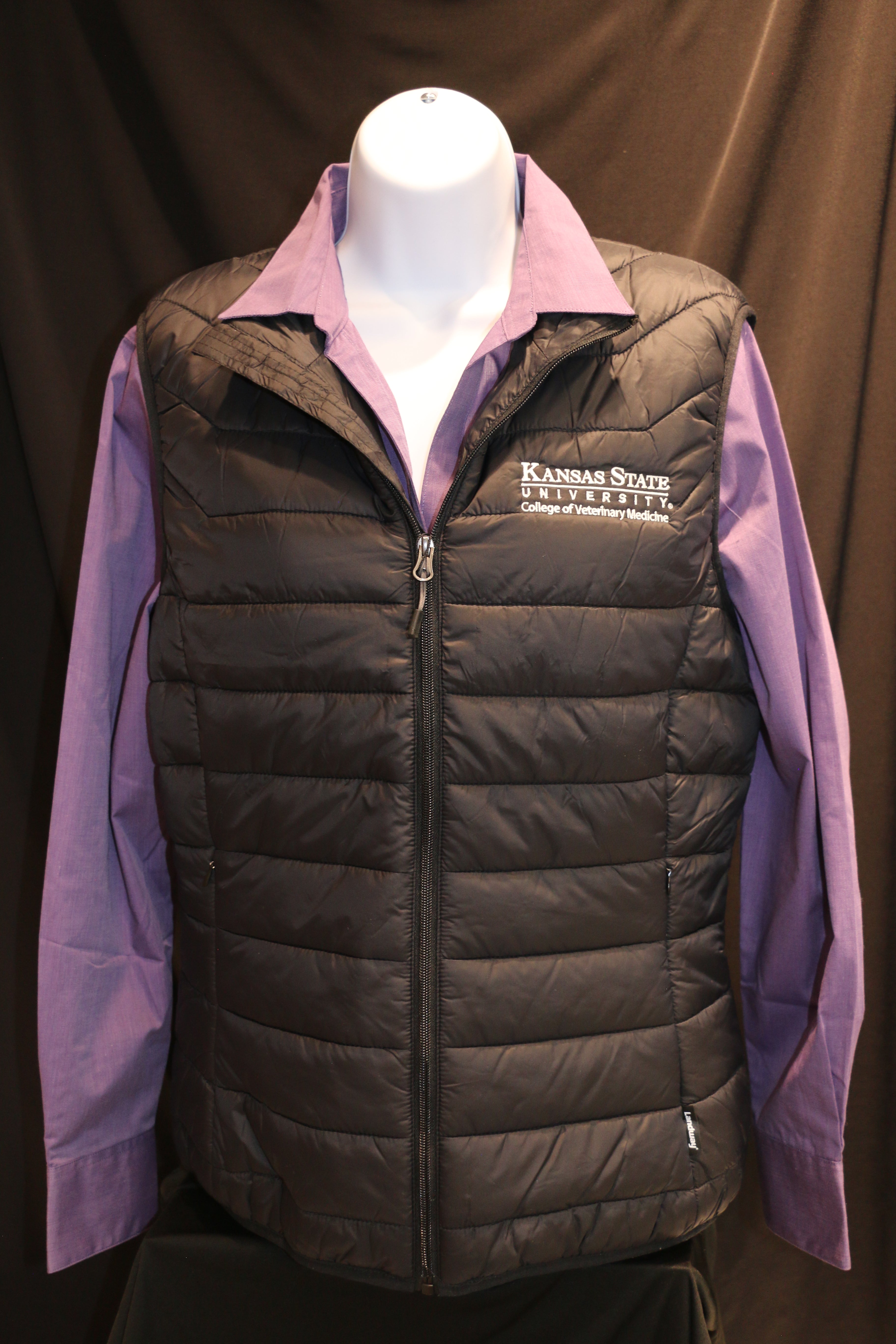 A black quilted puffer vest with zippered pockets, white K-State logo, and a stand-up collar unzipped on a mannequin.