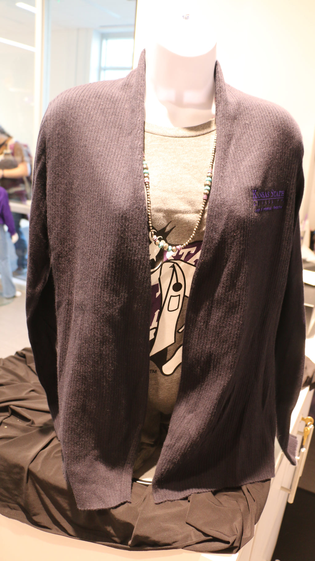 A lady's port authority ribbed black cardigan with purple "Kansas State University College of Veterinary Medicine" embroidered on the upper left chest area. 