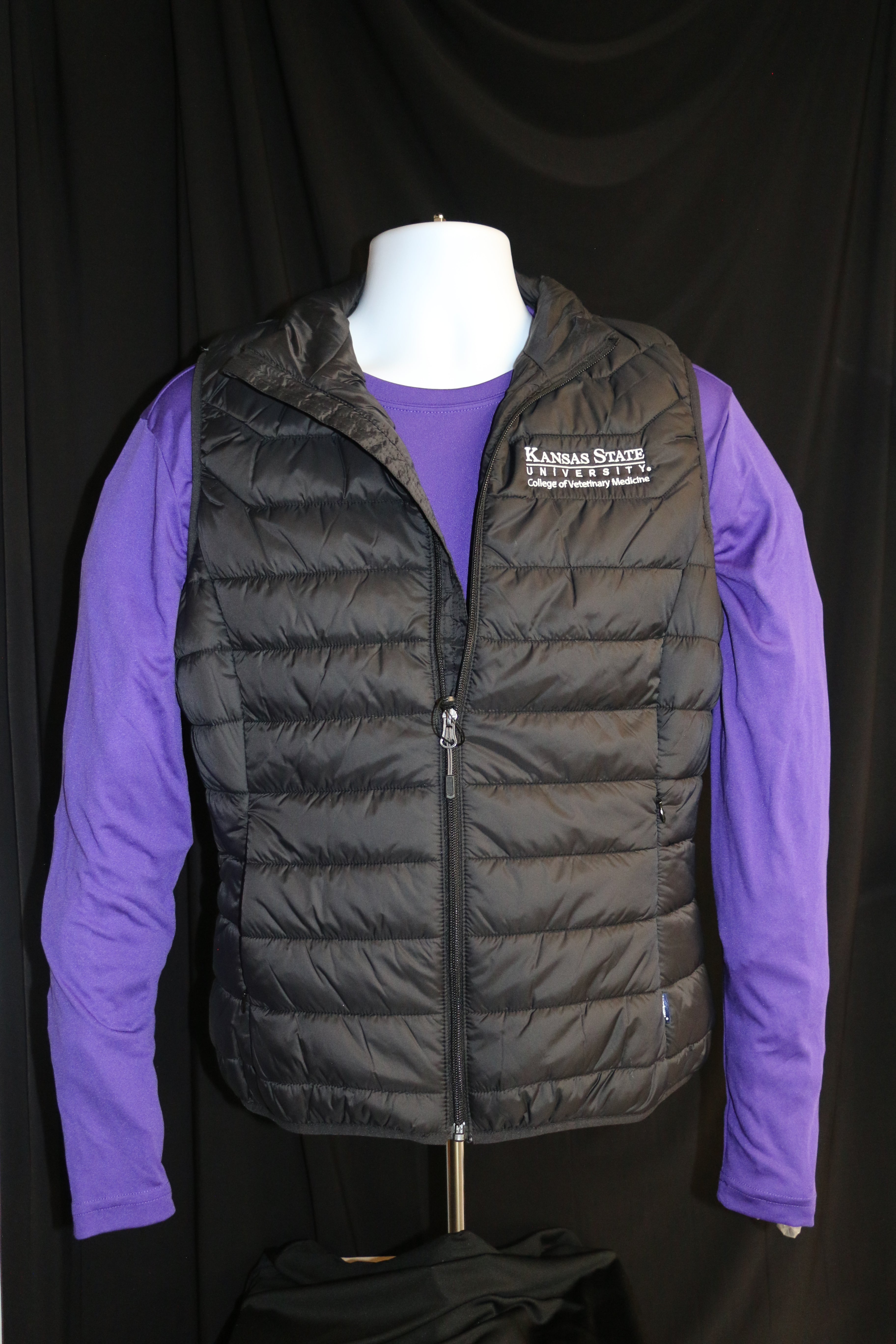 A mannequin wearing a black quilted puffer vest with zippered pockets, white K-State logo, and a stand-up collar, paired with a purple shirt.