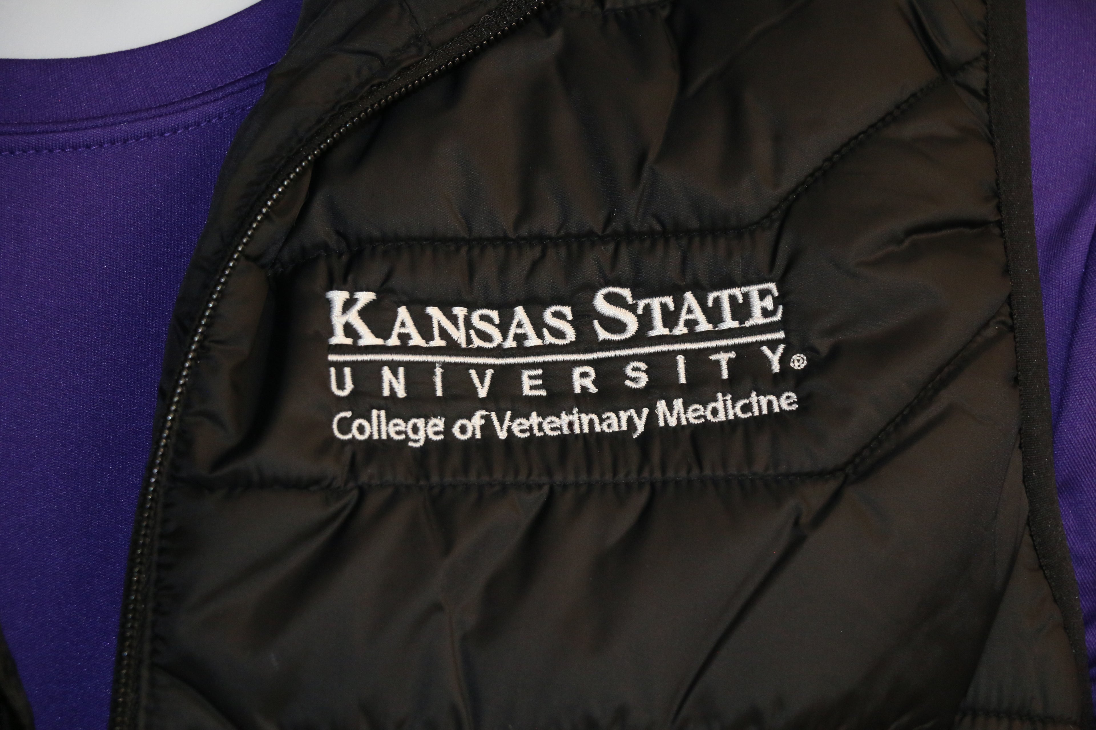 A close-up of the white "Kansas State University College of Veterinary Medicine" logo on the upper left side of the puffer vest. 