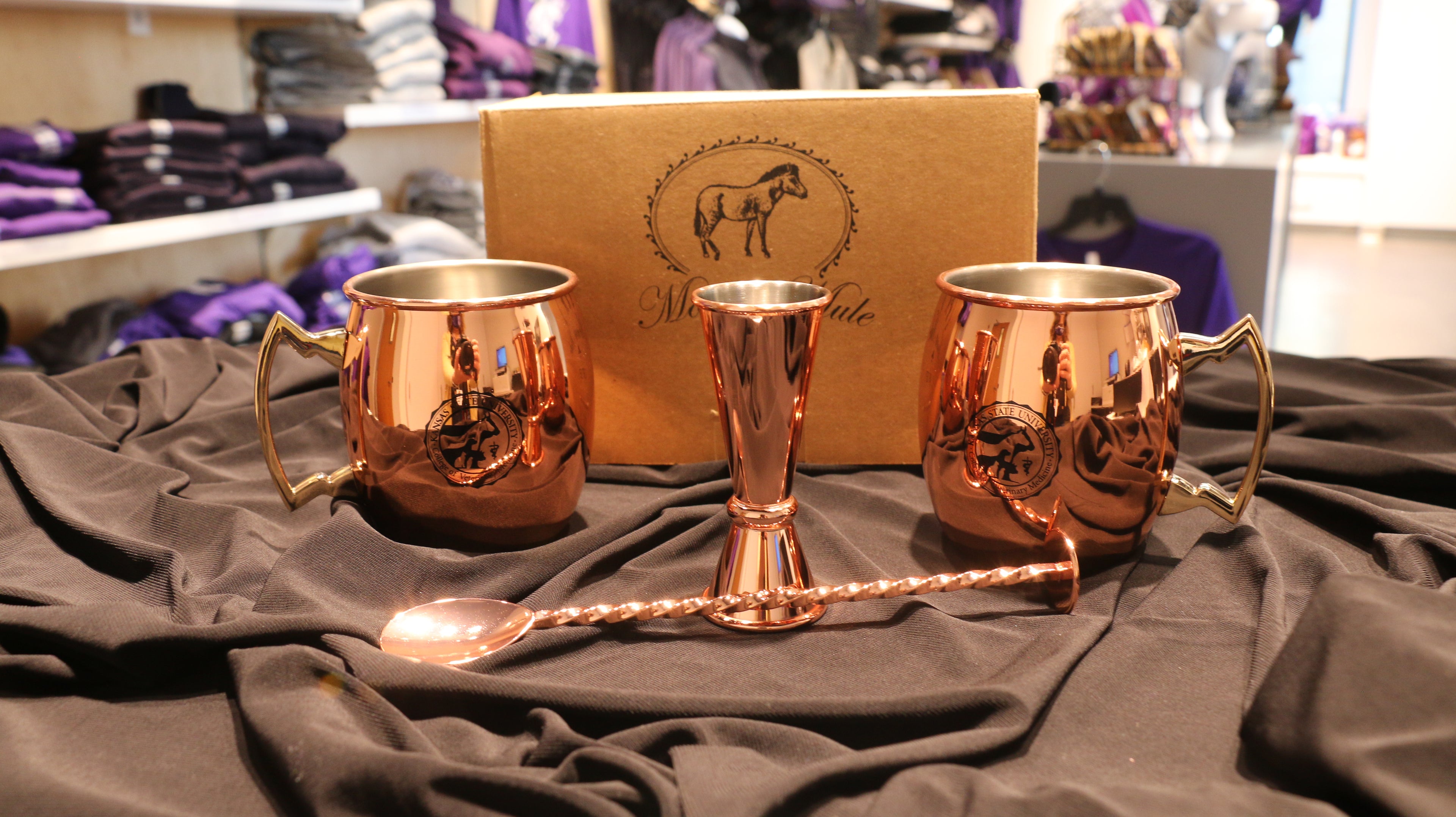 A set of two copper mugs with the CVM seal in black, a bar spoon, and a measuring jigger on a black fabric surface with a box in the background that has 'Moscow Mule' printed on it.