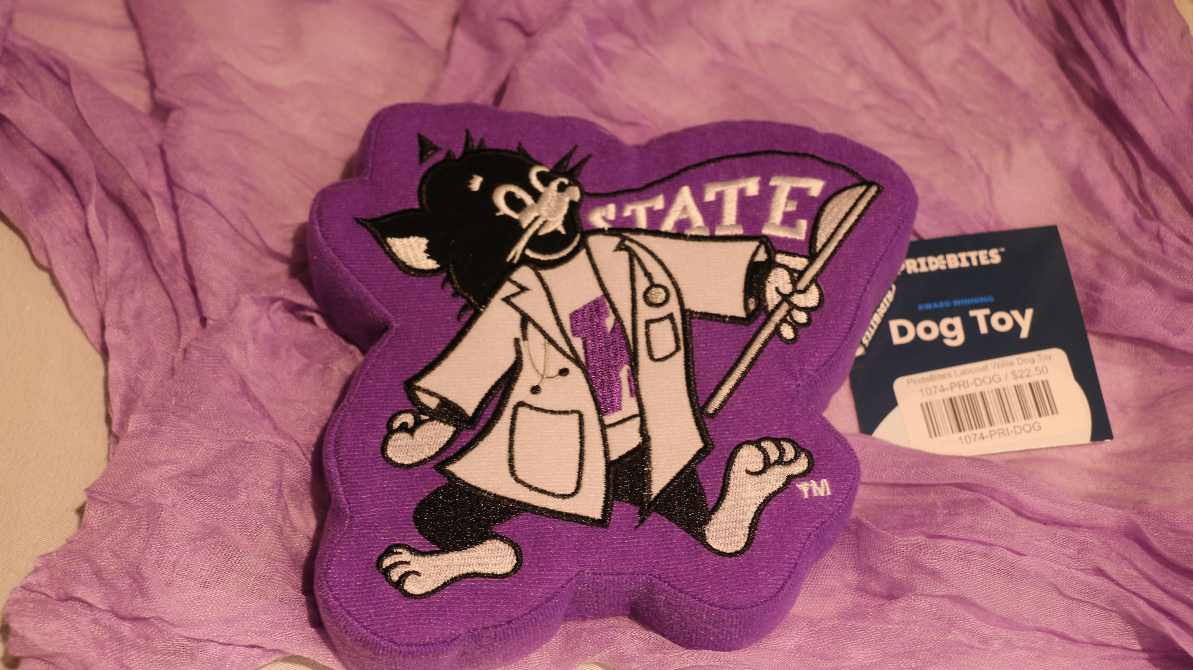 A purple dog toy shaped like Labcoat Willie, with a squeaker inside. The toy is placed on a purple surface, and there is a blue and white tag attached to it.