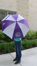 A person standing outdoors holding a purple and white striped umbrella with a white K-State College of Vet Med logo on one of the purple stripes. 