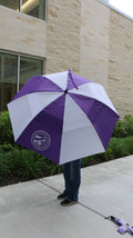 A person standing outdoors holding a purple and white striped umbrella with a white K-State College of Vet Med logo on one of the purple stripes. 