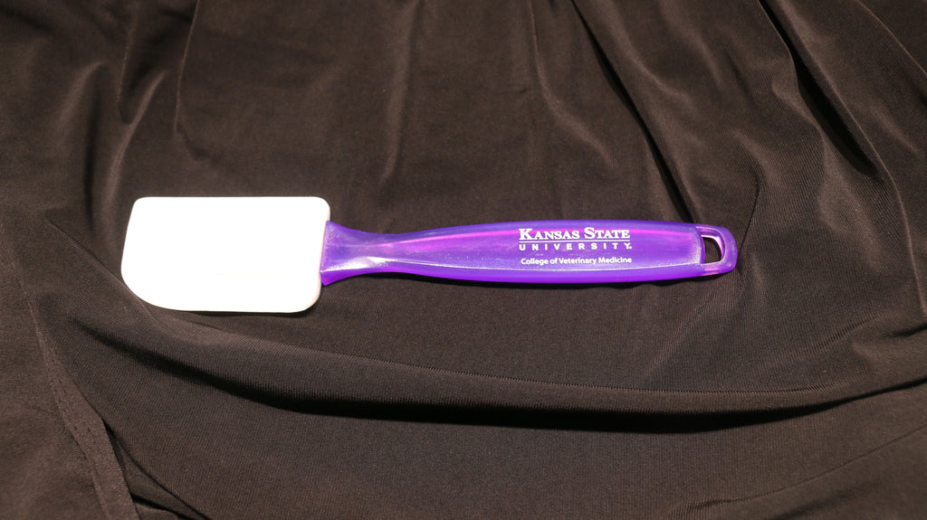 A purple silicone spatula with a translucent handle, white CVM logo, and lying on a black surface.