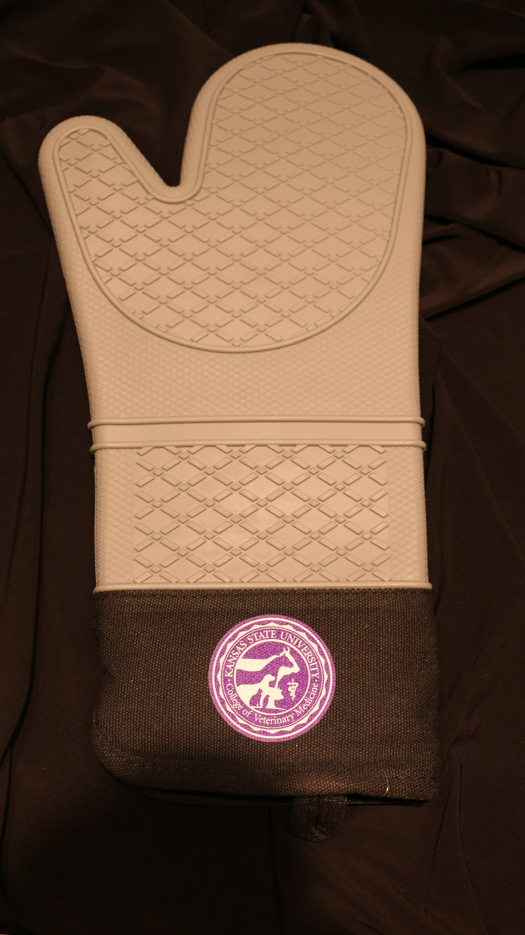 A silicone oven mitt with a light grey base color and black fabric cuff. On the center of the cuff on the mitt there is a white and purple CVM seal. 