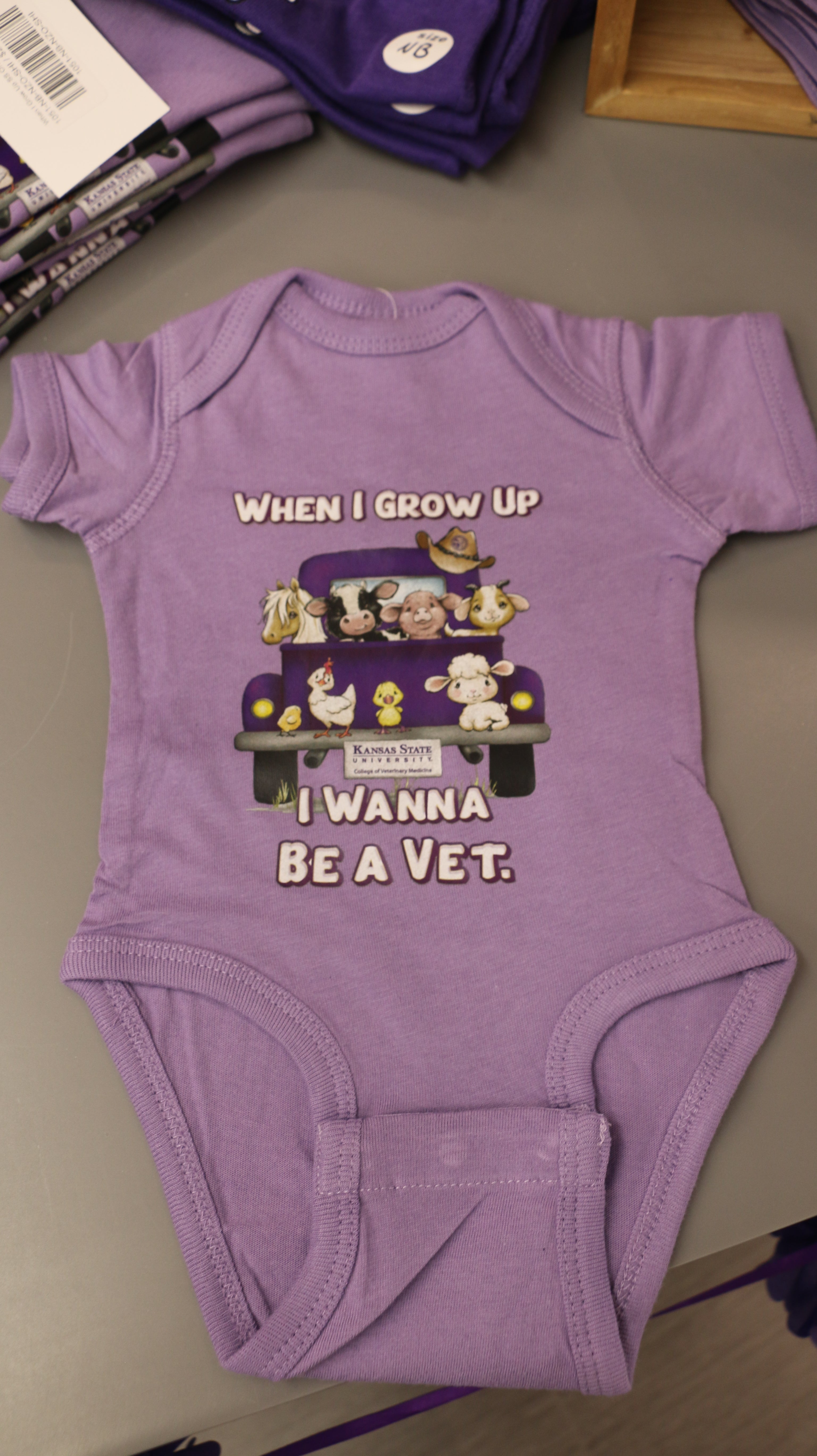 A lavender-colored short sleeve onesie with an illustration of different animals in the back of a truck and the text 'When I grow up I want to be a Vet'.